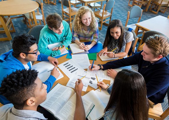 Diverse group of teen high school students are sitting around round table in library. They are studying books and papers to prepare for a test. Caucasian, African American, Hispanic, and Indian teen boys and girls are smiling while working together on class project.